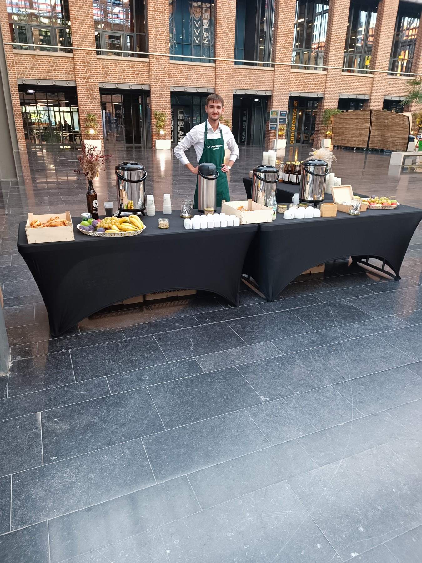 Un membre de l'équipe en tablier La Consignerie derrière le buffet du petit-déjeuner, sous la verrière de l'atrium
